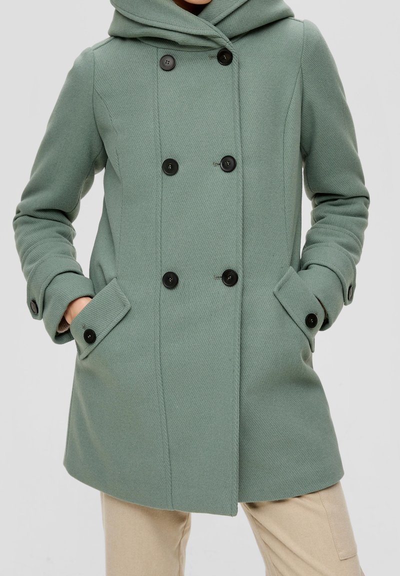 Manteau vert croisé avec boutons noirs, capuche et poches à rabat, porté avec les mains dans les poches, associé à un pantalon beige.