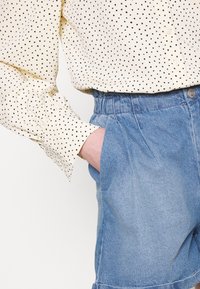 Vaaleankeltainen paita, jossa on mustia pilkkuja, pitkät hihat ja nappilista, yhdistettynä vaaleansinisiin farkkushortseihin, joissa on joustava vyötärö.