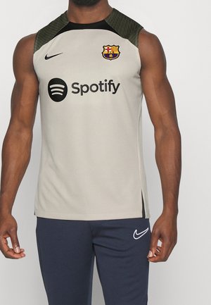 Mężczyzna noszący beżowy, bezrękawnikowy top sportowy z logo FC Barcelona i Spotify, zestawiony z granatowymi sportowymi spodniami Nike.