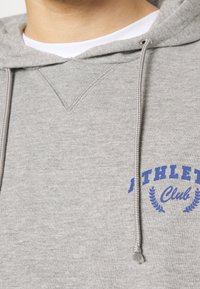Šedá mikina s kapucí z měkké látky, s přední kapsou ve stylu klokánka, kapucí se stahovací šňůrkou a modrým nápisem "Athletic Club" s vavřínovými akcenty.