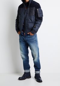 Hombre vestido con una chaqueta acolchada azul marino, jeans azules con puños, botas de cordones azul marino, con las manos en los bolsillos, de pie contra un fondo blanco.