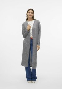 Grå lång kavaj med öppen framsida, tillverkad av mjukt stickat material, kombinerad med en vit tryckt topp och vida blå jeans.