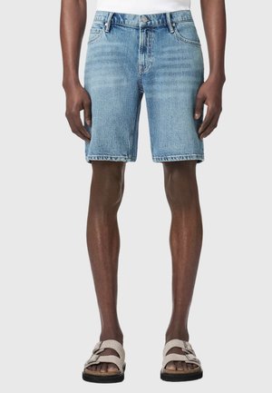 Mand iført lyseblå denimshorts og beige sandaler, stående med afslappede hænder langs siden mod en ensfarvet baggrund.