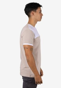 Camiseta beige con panel blanco en el hombro y detalles en las mangas. La tela es suave con un diseño ajustado. Detalle impreso notable en la parte frontal.