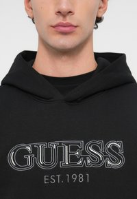 Zbliżenie na osobę noszącą czarną bluzę z kapturem, na której piersi wyszyto biały napis "GUESS EST. 1981".