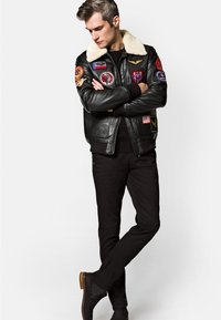 Czarna skórzana kurtka bomber z puszystym białym kołnierzem, ozdobiona różnorodnymi kolorowymi naszywkami, zestawiona z czarnymi spodniami i brązowym obuwiem za kostkę.