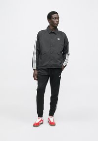 Črna športna jakna z belimi črtami na rokavih; črne hlače; rdeče-bele superge; minimalističen dizajn z logotipnimi poudarki.