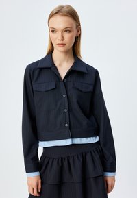 Giacca cropped blu navy con righe verticali, due tasche sul petto, chiusura con bottoni e accentuazione azzurro chiaro all'orlo. Tessuto morbido, design strutturato.