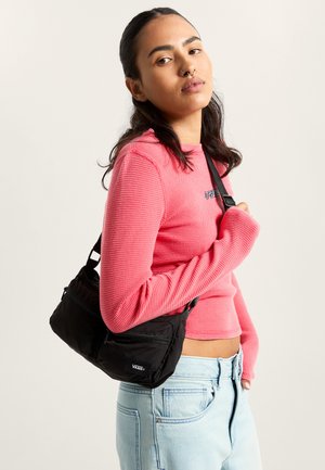 Sac bandoulière en nylon noir avec deux compartiments zippés, sangle ajustable et logo Vans. Porté avec un haut rose à manches longues.
