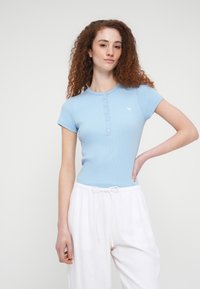 Ανοιχτό μπλε ριμπ κοντομάνικο μπλουζάκι με κουμπωτό λαιμό και μικρό λογότυπο, συνδυασμένο με λευκό χαλαρό παντελόνι. Λεία υφή και εφαρμοστή γραμμή.