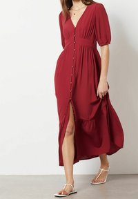 Žena v midi šatech s hlubokým červeným odstínem, knoflíkovým zapínáním, rozparkem po kolena a nabíranými rukávy, v bílé strappy ploché sandály, prochází uvnitř.