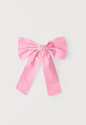 Ροζ υφασμάτινο φιόγκος με δύο μακριές ουρές και ένα μικρό χρυσό μενταγιόν στέμματος στο κέντρο, παρουσιασμένο σε απλό ανοιχτό φόντο.