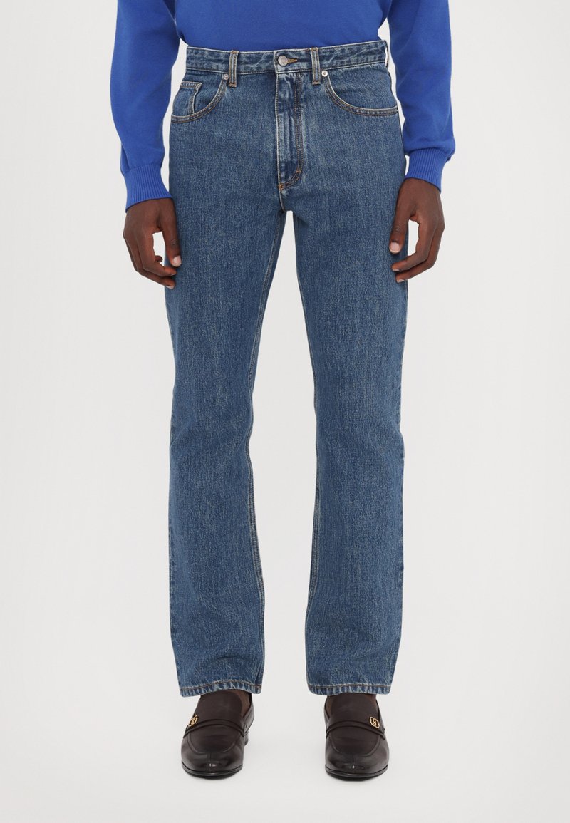 Blue denim jeans with a straight-leg fit, five pockets, and contrasting stitching. Worn with a blue sweater and brown shoes.