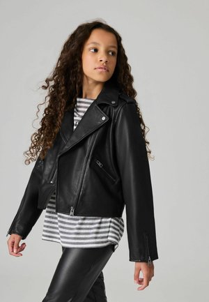 Zwarte leren motorjas met ritszakken, slim fit en brede revers, gedragen over een grijs-wit gestreept shirt en zwarte broek.