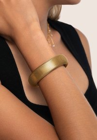 Mujer con una pulsera de oro texturizada en su muñeca y una blusa sin mangas negra, mostrada desde el cuello hasta el brazo contra un fondo neutral.