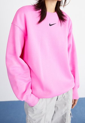 Osoba nosící oversize jasně růžovou mikinu Nike a světle šedé cargo kalhoty, s rukou v kapse, na bílém a modrém pozadí.
