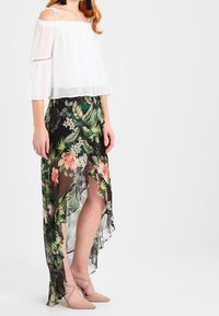 Weiße Bluse mit schulterfreier Optik aus transparentem Stoff; schwarzer Rock mit floralem Muster und asymmetrischem Saum in Grün- und Rosatönen; nudefarbene Schuhe.