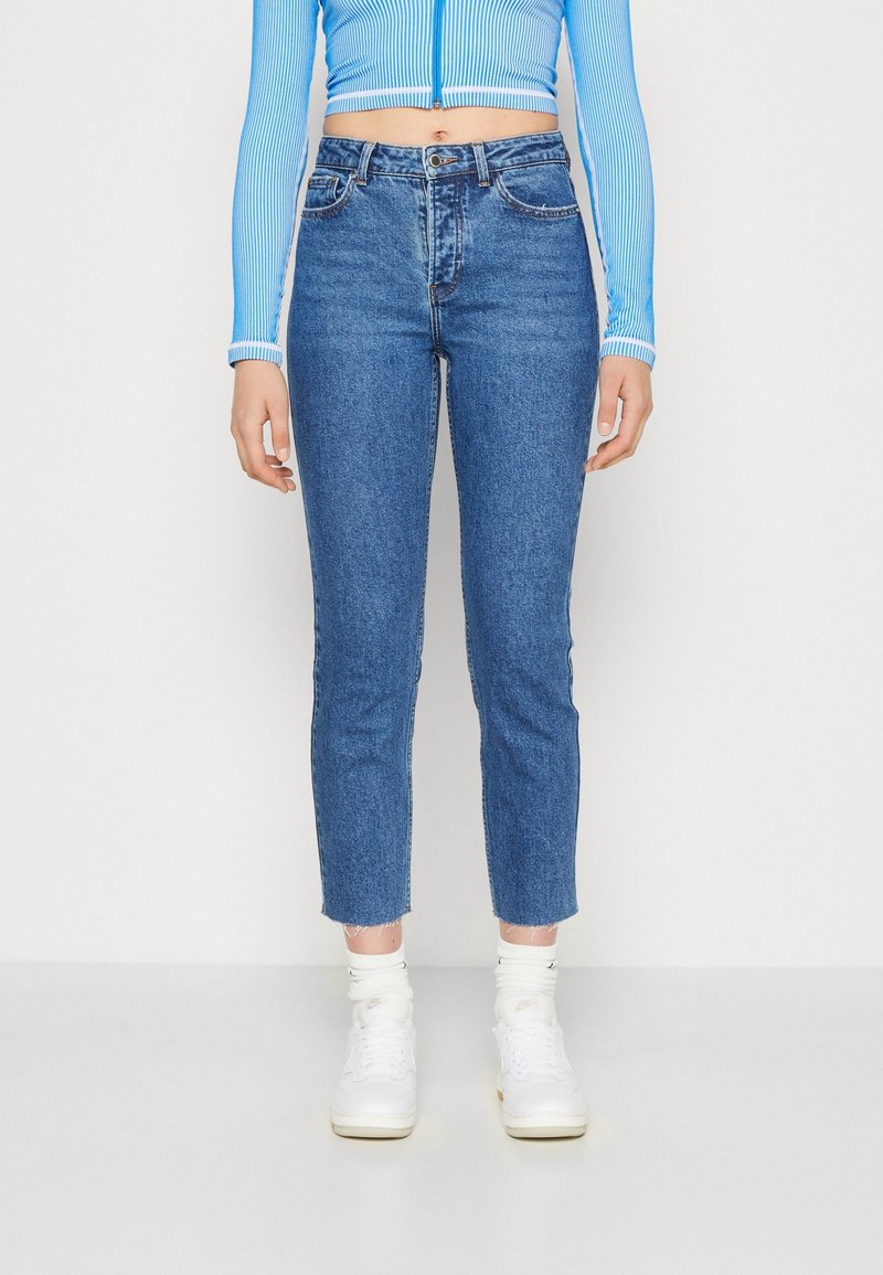 Blå denimjeans med smal passform, fransade fållar och femficksdesign, kombinerat med vita sneakers och krämfärgade strumpor.