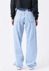 Light blue wide-leg jeans, denim fabric, back pockets. Features a black brand label at the waist. Casual fit with a relaxed silhouette.