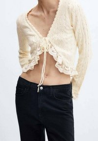 Kremowy, długi sweter-kardigan z dzianiny o krótkim kroju, z dekoltem w serek, koronkową lamówką i zapięciem na sznurek. W połączeniu z czarnymi, wysokim stanem dżinsami.