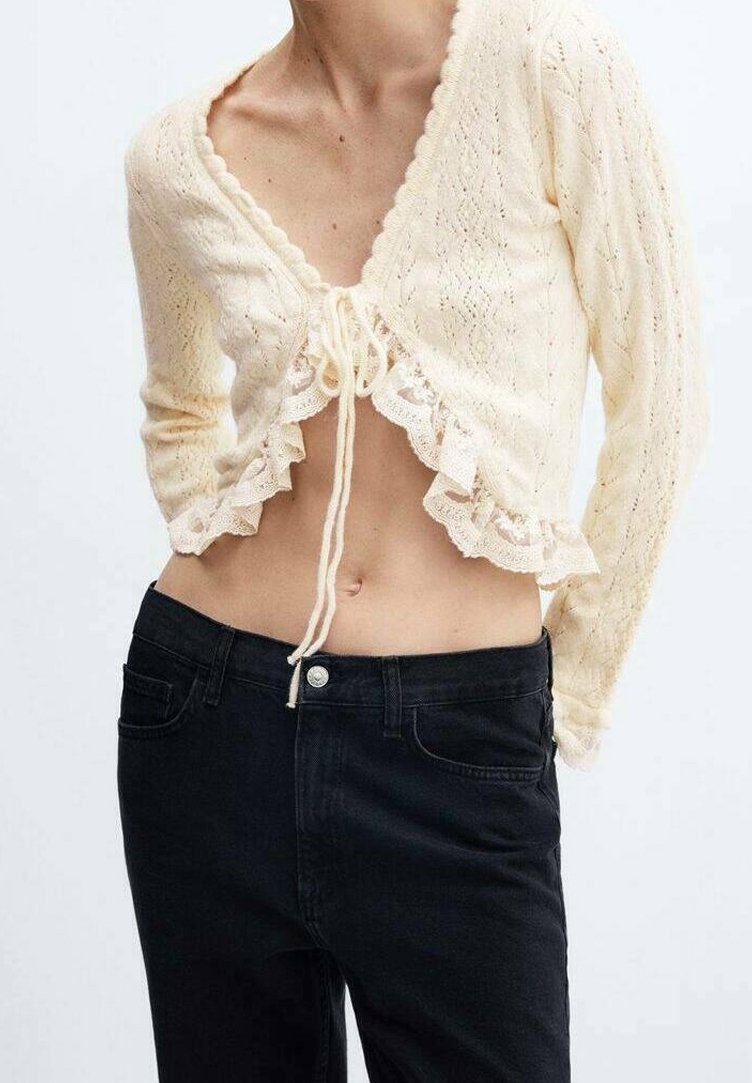 Kremowy, długi sweter-kardigan z dzianiny o krótkim kroju, z dekoltem w serek, koronkową lamówką i zapięciem na sznurek. W połączeniu z czarnymi, wysokim stanem dżinsami.