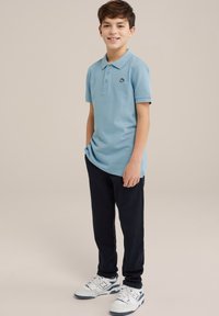 Gaiši zila īsām piedurknēm polo krekls ar apkakli un izšūtu logotipu, apvienojot ar tumšzilu biksēm un baltajām kedām ar zilu akcentu.