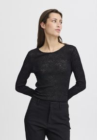 Top nero a maniche lunghe con texture in pizzo floreale, scollo rotondo, design aderente, abbinato a pantaloni neri sartoriali.