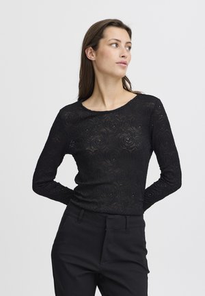 Top nero a maniche lunghe con texture in pizzo floreale, scollo rotondo, design aderente, abbinato a pantaloni neri sartoriali.
