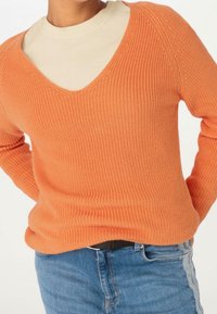 Pomarańczowy sweter dziany z dekoltem w kształcie litery V i prążkowaną strukturą, założony na kremowy top z wysokim kołnierzem. Noszony z niebieskimi dżinsami z bocznym paskiem.