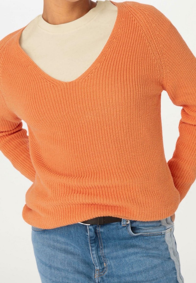 Pomarańczowy sweter dziany z dekoltem w kształcie litery V i prążkowaną strukturą, założony na kremowy top z wysokim kołnierzem. Noszony z niebieskimi dżinsami z bocznym paskiem.