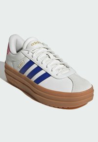 Biały tenisówka z niebieskimi paskami i różowym akcentem. Posiada teksturowaną cholewkę, okrągły nosek oraz grubą podeszwę z gumy w kolorze brązowym. Złote logo jako detal.