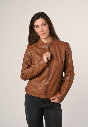 Frau mit langen dunklen Haaren, die eine braune Lederjacke und dunkle Jeans trägt, hält den Reißverschluss der Jacke nahe ihrer Brust vor einem schlichten Hintergrund.