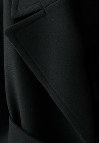 Zwarte wollen blazer met een textuurafwerking, scherpe revers en een getailleerd silhouet met zichtbare stikdetails.