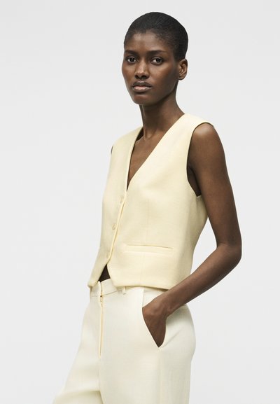 Γυναίκα που φοράει αμάνικο ελεφαντόδοντο γιλέκο και ασορτί ψηλόμεσο παντελόνι, στέκεται με το ένα χέρι στην τσέπη μπροστά σε απλό φόντο.