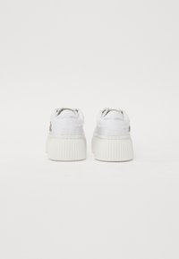 Sneaker bianche con suola texturizzata e scanalata, dotate di una parte superiore liscia e un branding discreto sul retro. I lacci sono ben in vista.