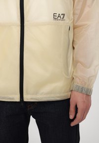 Lightweight beige jacket made of semi-transparent material with black zipper, elastic cuffs, and EA7 logo on the chest.