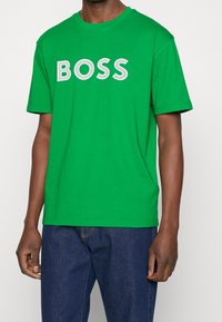 Zelené bavlněné tričko s velkým bílým perforovaným nápisem "BOSS". Krátké rukávy a kulatý výstřih.