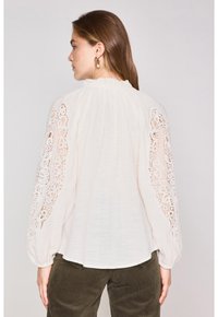 Blusa color beige chiaro con maniche lunghe e voluminose, caratterizzata da dettagli di pizzo intricati lungo le spalle e un colletto arricciato.
