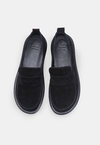 O pereche de mocasini negri din suede cu tălpi din piele și o talpă din cauciuc cusută, văzuți de sus pe un fundal alb.