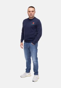 Felpa navy con piccolo graphic di una mela, jeans blu e sneaker grigio chiaro. Vestibilità casual con polsini e orlo a costine. Sfondo semplice.