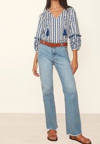 Modrá a bílá vzorovaná blůza s vycpanými rukávy, kterou doplňují světle modré kalhoty s vysokým pasem, hnědým opaskem a béžovými sandály.