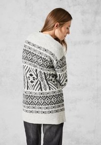 Lång, grå stickad cardigan med svarta geometriska mönster. Har rund halsringning, långa ärmar och en avslappnad passform. Slät textur.
