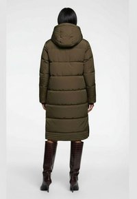 Olivgrön vadderad jacka med huva, utrustad med horisontella quiltade paneler och mitt på vaden-längd. Bärs med glänsande bruna knähöga stövlar.