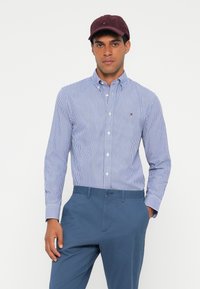 Blau-weiß gestreiftes Hemd mit Knopfleiste und Kragen, kombiniert mit blauen Hosen. Eine bordeauxfarbene Mütze sitzt auf dem Kopf des Models.