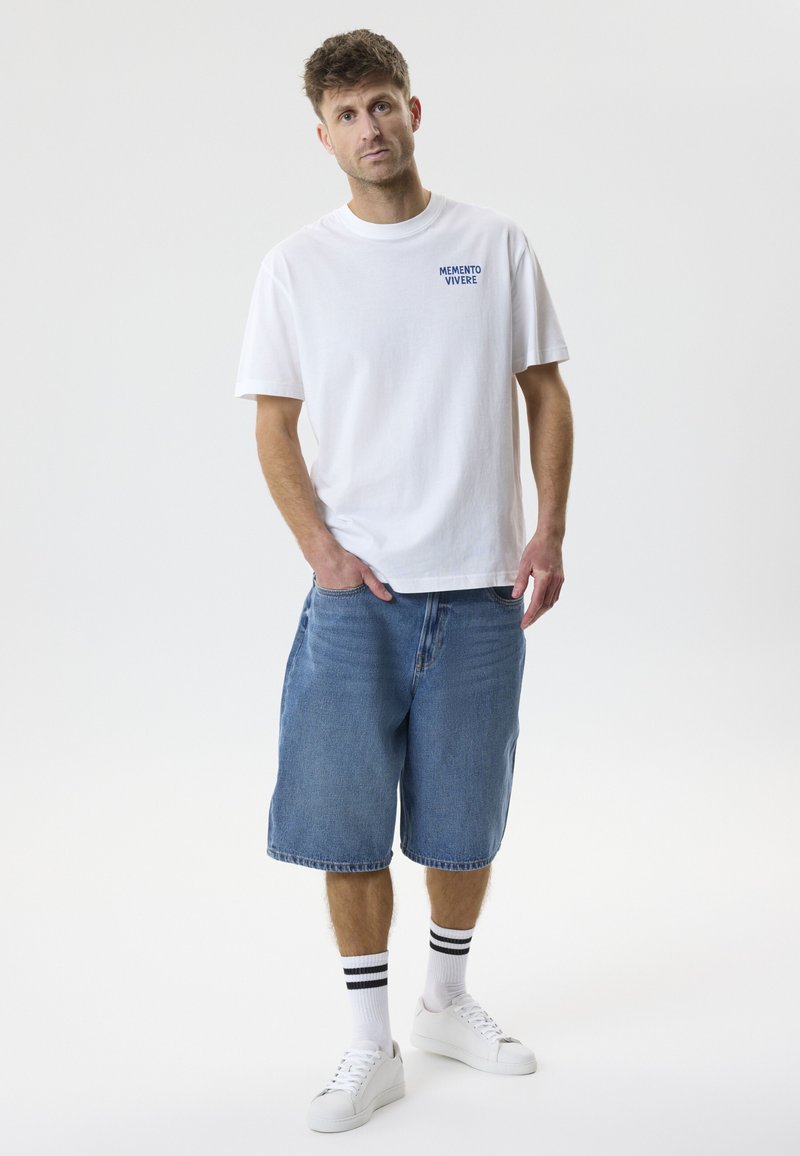 Jeune homme debout, les mains dans les poches, portant un T-shirt blanc avec le texte « Memento Vivere », un short en jean bleu, des chaussettes rayées et des baskets blanches.