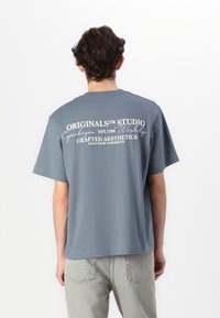 Grå bomullströja med korta ärmar, med vit text på baksidan: "Originalism Studio Köpenhamn Est. 1990" och "Crafted Aesthetics."