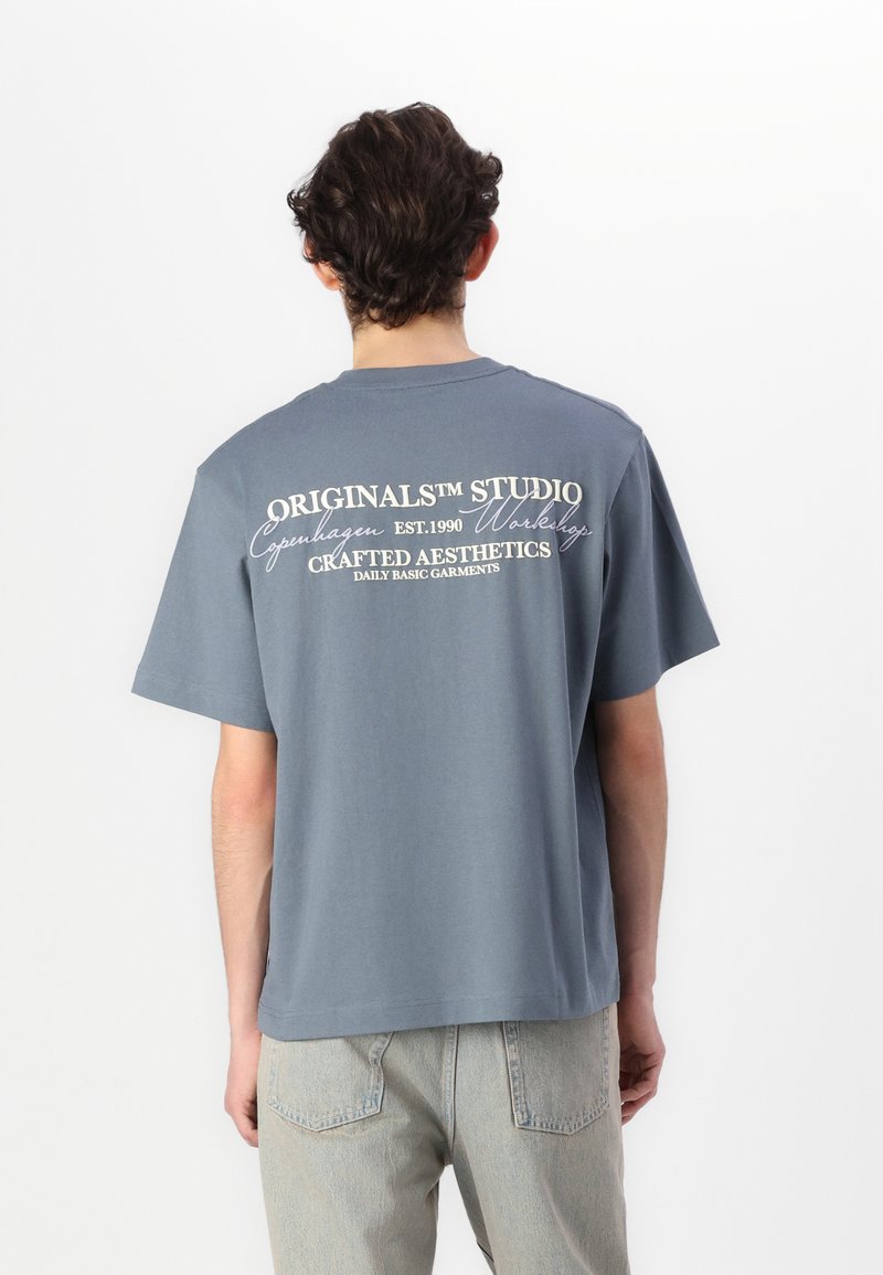 Grå bomulds-t-shirt med korte ærmer, prydet med hvid tekst på ryggen: "Originalism Studio Copenhagen Est. 1990" og "Crafted Aesthetics."