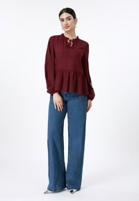 Μπορντό τοπ με βολάν στο λαιμό και λεπτομέρεια δέσιμου, σε συνδυασμό με μπλε τζιν παντελόνι με φαρδιά μπατζάκια και λευκές μυτερές γόβες.