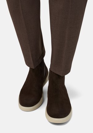 Ruske nubuck ankletossor med en lågprofil vit gummiklåsol. Passformen är snug och designad för avslappnad användning, visad i kombination med bruna byxor.