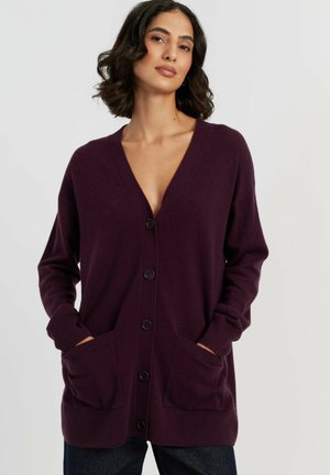 Vrouw draagt een diep paarse gebuttonde cardigan met zakken, handen in de zakken, en een donkere broek tegen een effen lichte achtergrond.